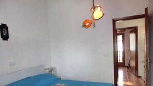 Un dormitorio con una colcha azul, una lámpara fijada a la pared y una puerta que da a otra habitación.