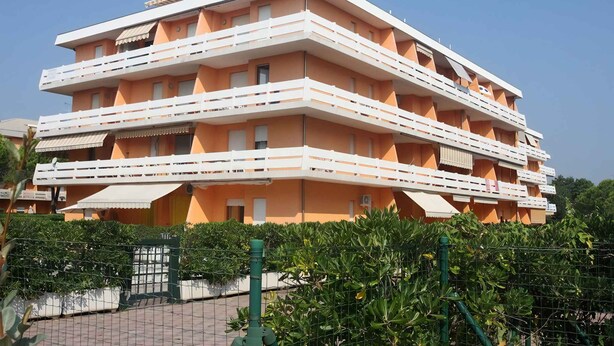 Un edificio de varios pisos con balcones, una entrada protegida por un portón y abundante vegetación alrededor.