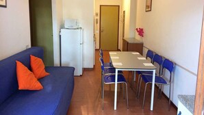 Un espacio habitable compacto con un sofá azul, cojines anaranjados, una mesa de comedor con sillas, un refrigerador y una pequeña zona de cocina.