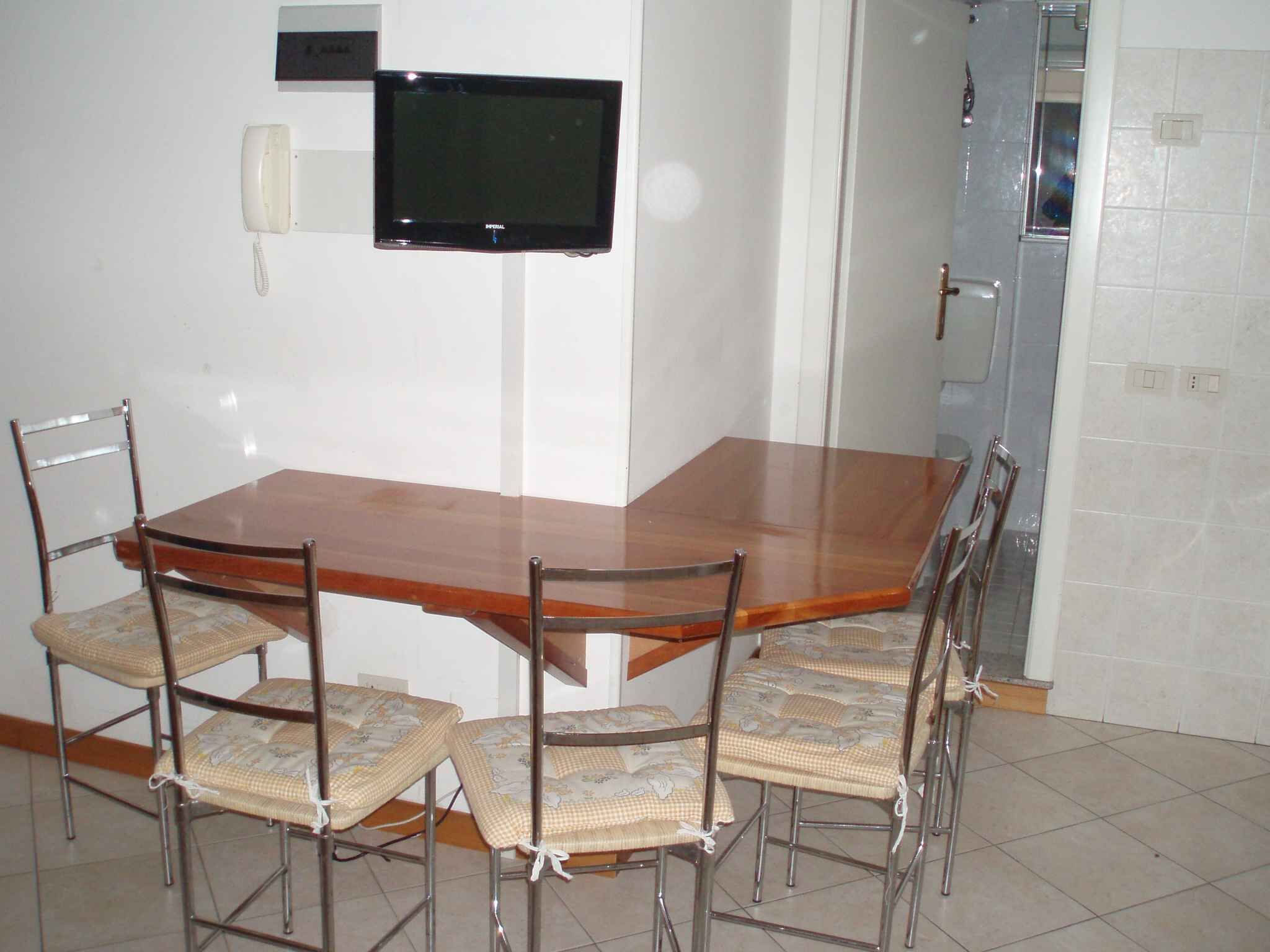 Un coin repas avec une table en bois et six chaises, un téléviseur fixé au mur, et une salle de bain visible au fond.