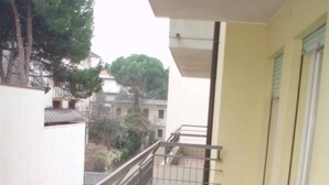 Un balcón estrecho con suelo de baldosas, barandilla de metal y vistas a edificios y árboles cercanos.