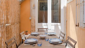Une table à manger dressée avec des assiettes, des verres et des couverts, entourée de chaises en bois, avec une fenêtre et une porte en arrière-plan.