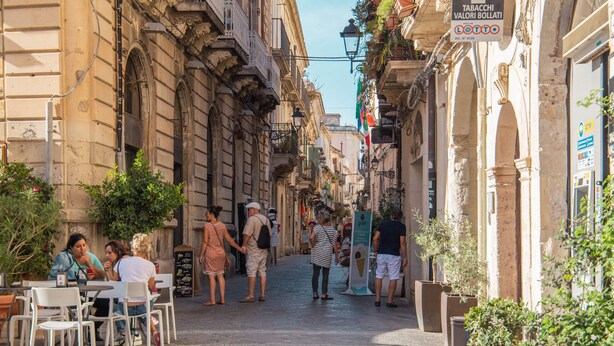 A narrow street with outdoor seating, potted plants, and historic buildings. - Casa Darsena with Sea View Balcony by Wonderful Italy (Siracusa)