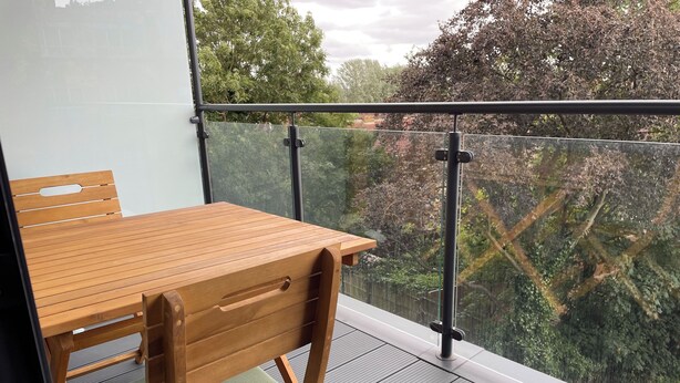 Een houten tafel en stoelen op een balkon met een glazen balustrade, met uitzicht op een bebost gebied.