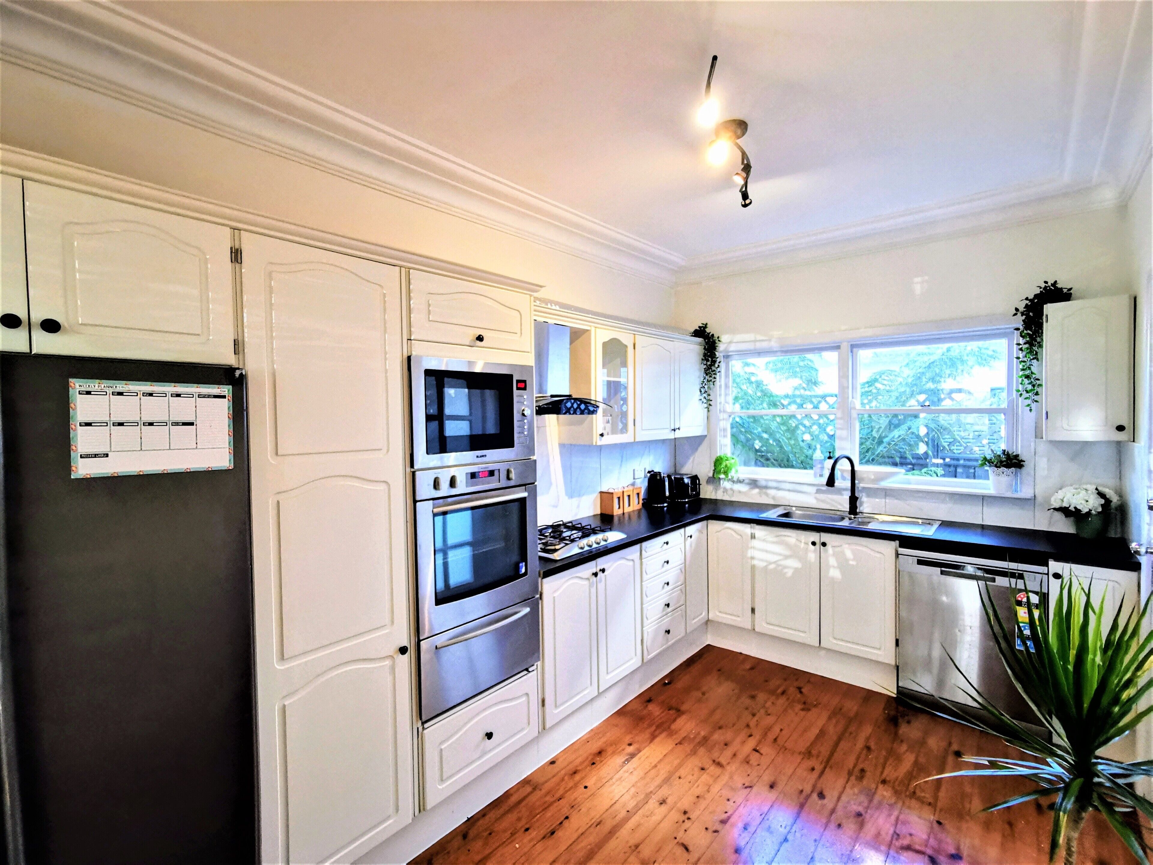 A kitchen with white cabinets, a black refrigerator, a microwave, an oven, a sink, and a window with a view of greenery outside.