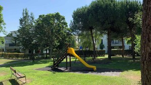Un parque infantil con un tobogán amarillo y columpios, rodeado de árboles y césped.