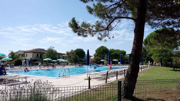 Un área de piscina con sillones de descanso y sombrillas, rodeada de árboles y edificios.