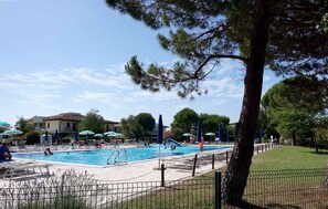 A swimming pool area with lounge chairs and umbrellas, surrounded by trees and buildings. - Holiday apartment with balcony and pool (Caorle)