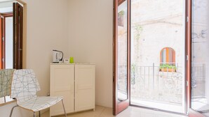 Et værelse med et hvidt skab, en stol med et geometrisk mønster og en skydedør, der fører til et udendørs område med en balkon og udsigt til en bygning med et vindue.