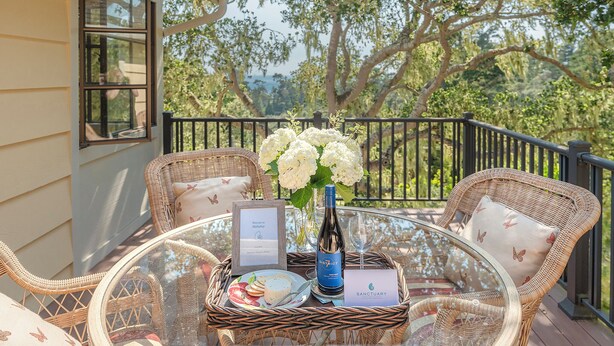 A wicker table with a glass top, a bottle of wine, a framed menu, and a plate of food on a balcony with a view of trees. - 3903 Mabuhay - Walk to Downtown and the Beach (Carmel)