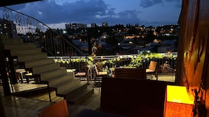 A terrace with a staircase, outdoor seating, and a view of a hillside town at dusk. - Hotel Piano (Tbilisi)