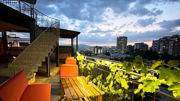 A rooftop terrace with wooden tables and orange cushions, a staircase, and a view of a cityscape under a cloudy sky. - Hotel Piano (Tbilisi)