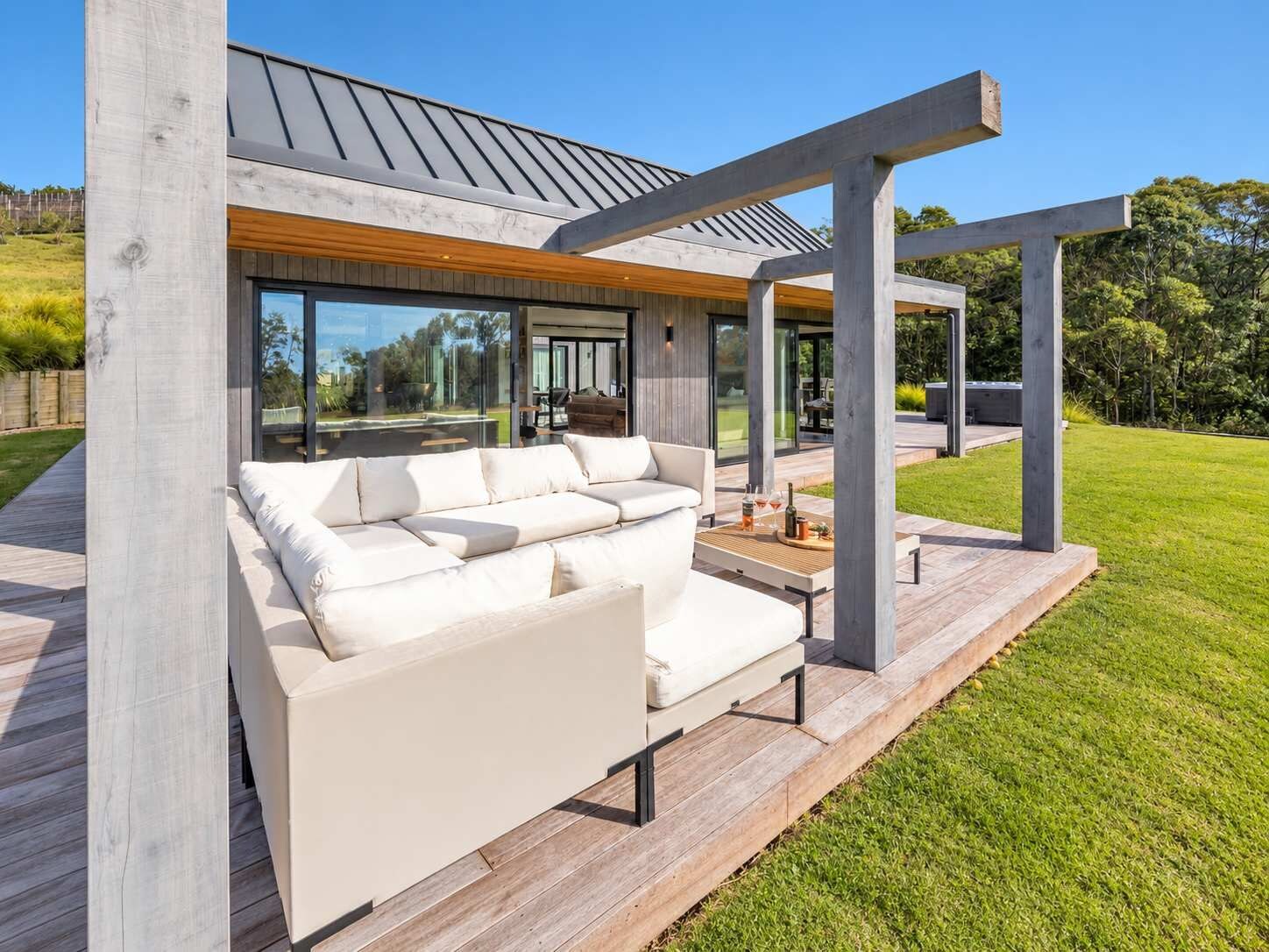A modern outdoor living area with a white sofa and coffee table, wooden deck, and a pergola structure.