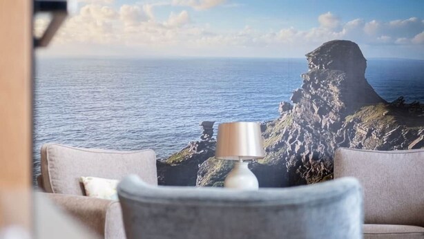A room with a view of the ocean and a rocky cliff. There is a lamp on a table and two chairs.