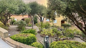 Un jardín ajardinado con varios árboles y arbustos, un sendero de piedra y un edificio con fachada amarilla al fondo.