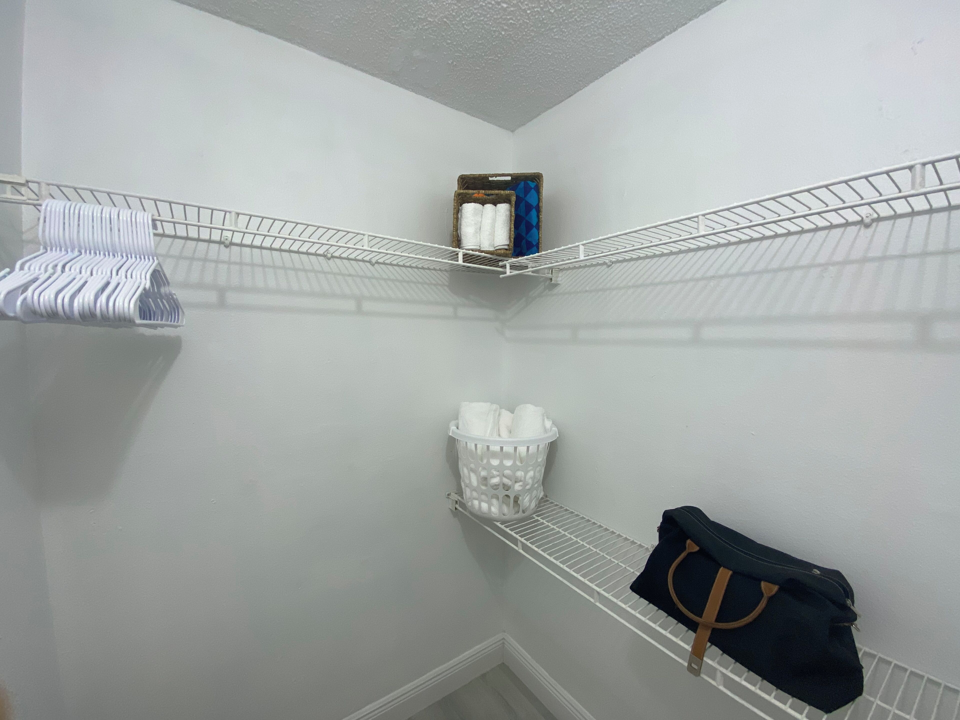 Eine Ecke eines Raumes mit weißen Wänden, einer weißen Decke und einem weißen Boden. Eine weiße Drahtablage mit einem weißen Korb und einem weißen Kunststoffbehälter darauf.