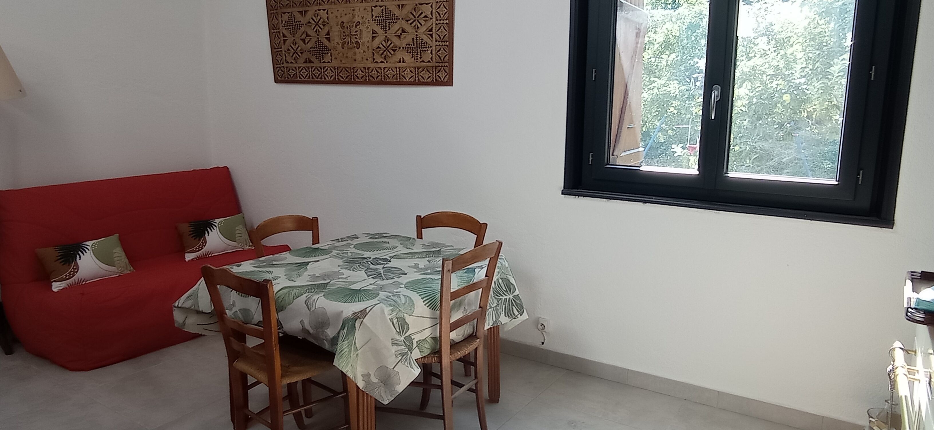 Un coin repas avec une table recouverte d’une nappe à motifs floraux, entourée de chaises en bois, d’un canapé rouge agrémenté de coussins à motifs et d’une fenêtre donnant sur un paysage verdoyant.