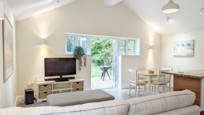 A modern living room with a flat-screen TV, a sofa, a dining table with chairs, and a kitchen area. - Millside, Bosham -  a bungalow that sleeps 4 guests  in 1 bedroom (Bosham)