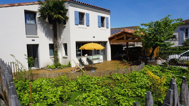 Ein modernes, zweistöckiges Haus mit einer Holzterrasse, einem gelben Sonnenschirm und einem umzäunten Garten.