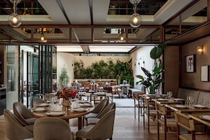 Un restaurante con decoración moderna, mesas y sillas de madera, una planta grande en maceta y vista del área de mesas al aire libre.