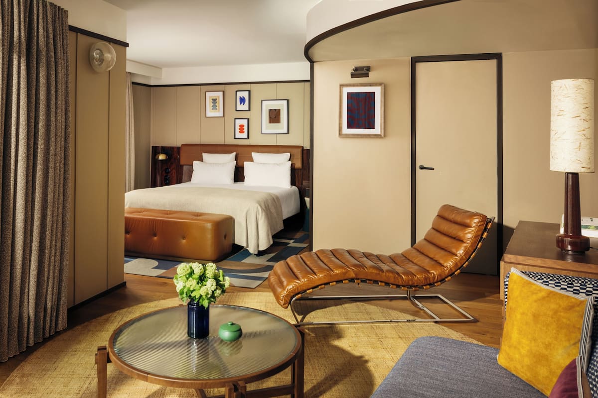 Habitación de hotel con cama, una butaca, una mesita con un florero y una lámpara.