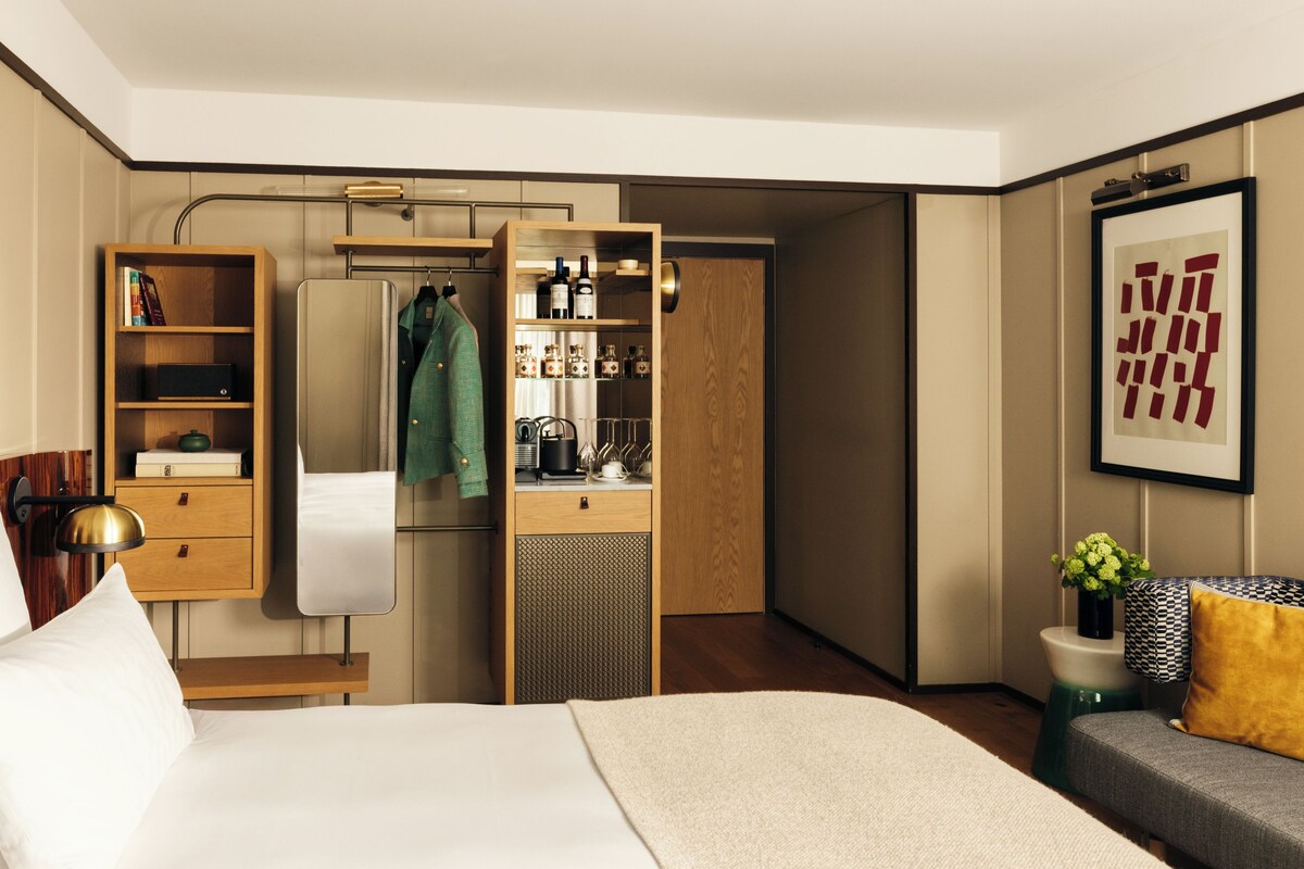 Una habitación de hotel moderna con una cama, un armario, un tocador con espejo y una zona de estar con un sofá y una mesita.