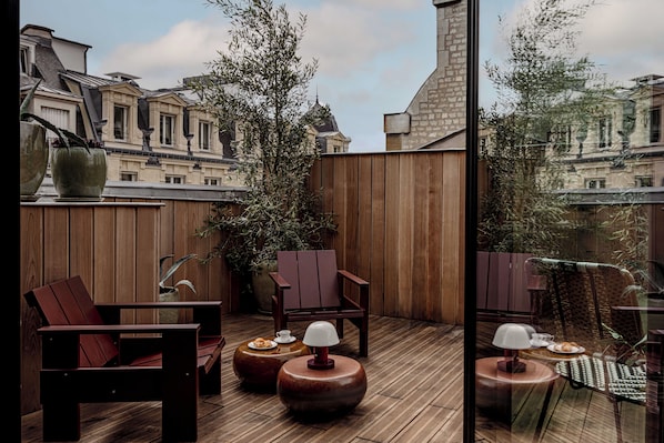 Una terraza de madera con zona de estar, que incluye sillas y mesas redondas, con vista a un paisaje urbano.