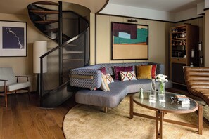 Ein modernes Wohnzimmer mit einer geschwungenen Treppe, einem Ecksofa mit gemusterten Kissen, einem Glastisch und einem Ledersessel.