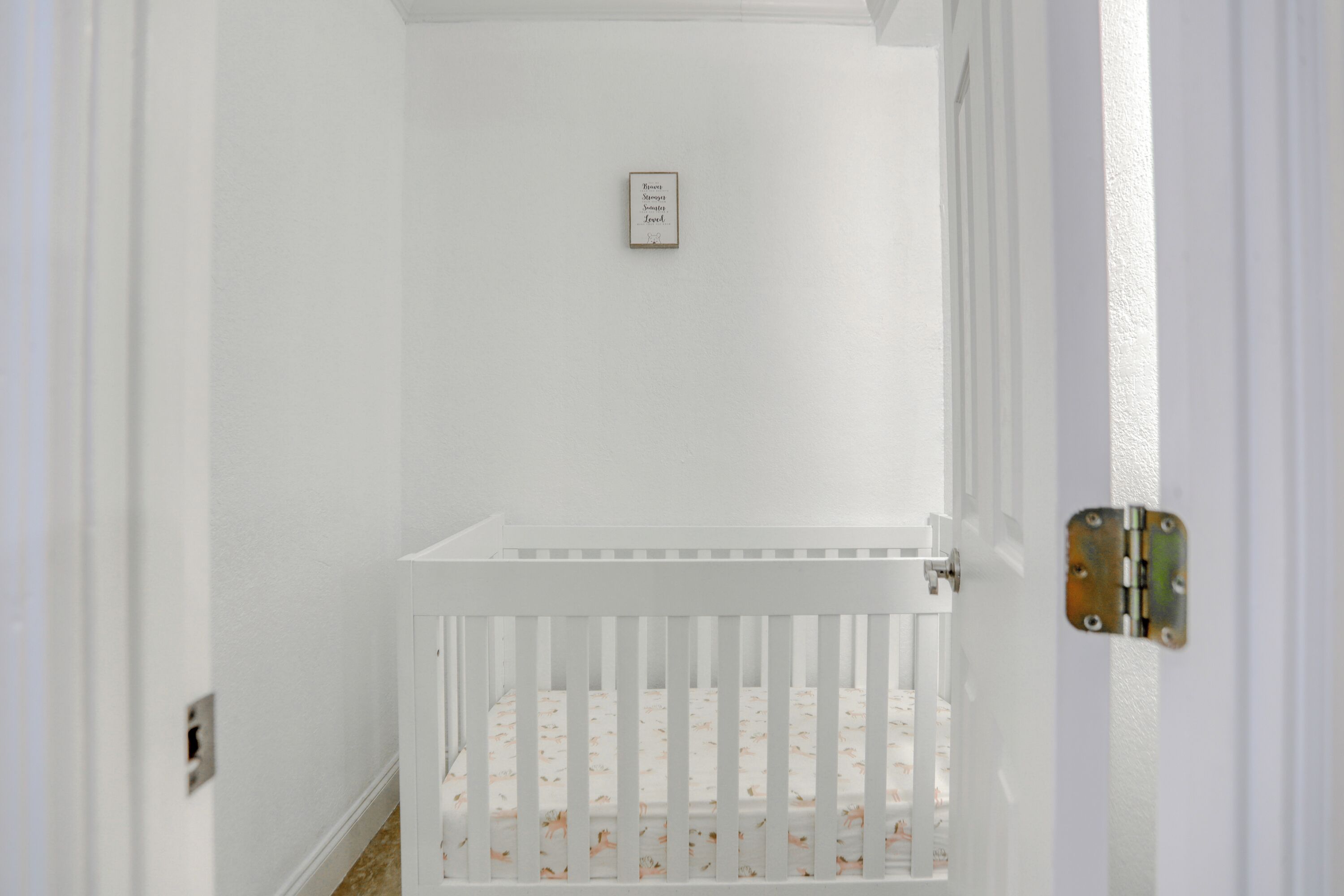 A white crib with a floral mattress in a room with white walls and a door with a metal latch.