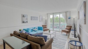 Una sala de estar moderna con un sofá azul, sillones marrones, una mesa de centro y un televisor. Hay cuadros enmarcados en las paredes y una vista al exterior a través de la puerta de vidrio.