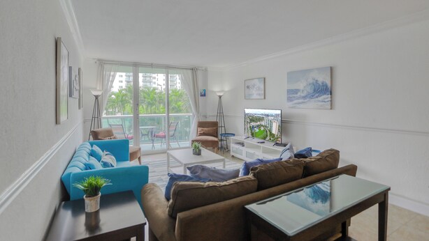 Una sala de estar moderna con un sofá azul, un sofá marrón y una mesa de centro de vidrio. Hay un balcón con vistas a palmeras y edificios.