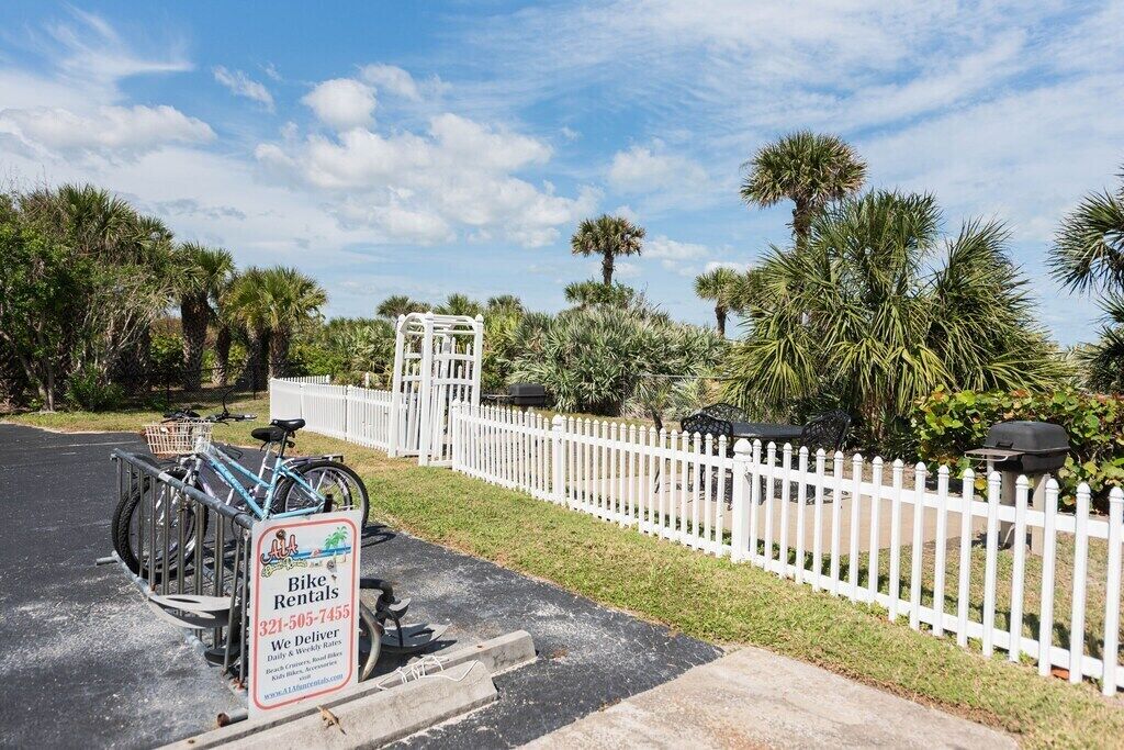 A bike rental sign in a parking lot with bicycles, a white picket fence, and palm trees in the background.