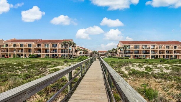 A wooden boardwalk leading to a row of buildings with multiple balconies, surrounded by grassy areas and sand dunes.