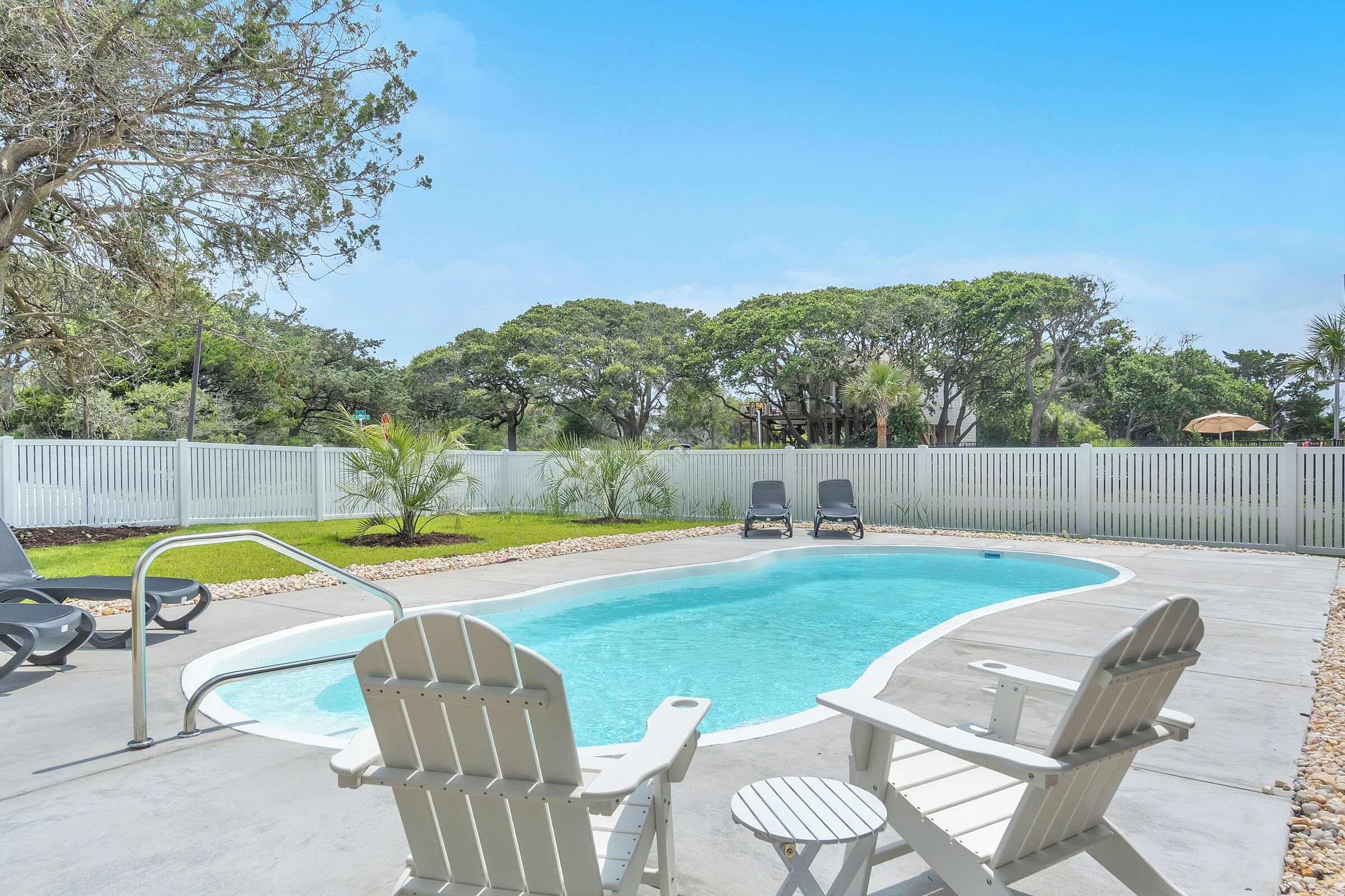 A backyard with a swimming pool, white Adirondack chairs, and a white fence.