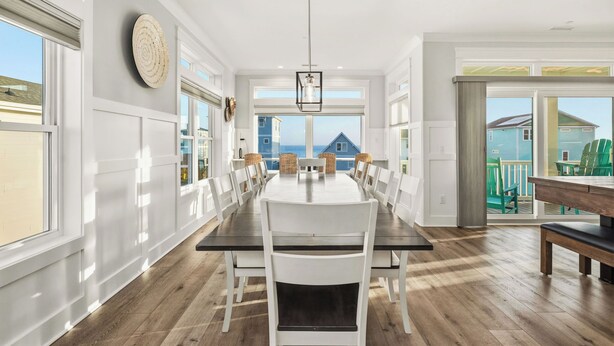 A dining area with a long table, white chairs, and a view of the ocean. - Slow M'Ocean: Large, Modern Home with a Pool, Elevator and Hot Tub! (Oak Island)