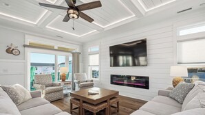 A modern living room with a ceiling fan, a flat-screen TV, a fireplace, and a large window. - Slow M'Ocean: Large, Modern Home with a Pool, Elevator and Hot Tub! (Oak Island)