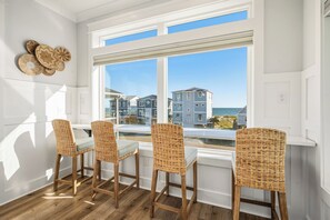 A room with a white interior, a large window with a view of buildings and the sea, and four wicker chairs with a small table. - Slow M'Ocean: Large, Modern Home with a Pool, Elevator and Hot Tub! (Oak Island)