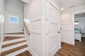 A staircase with wooden steps leading to a white door, a small window, and a view into a bedroom with a bed and nightstand. - Slow M'Ocean: Large, Modern Home with a Pool, Elevator and Hot Tub! (Oak Island)