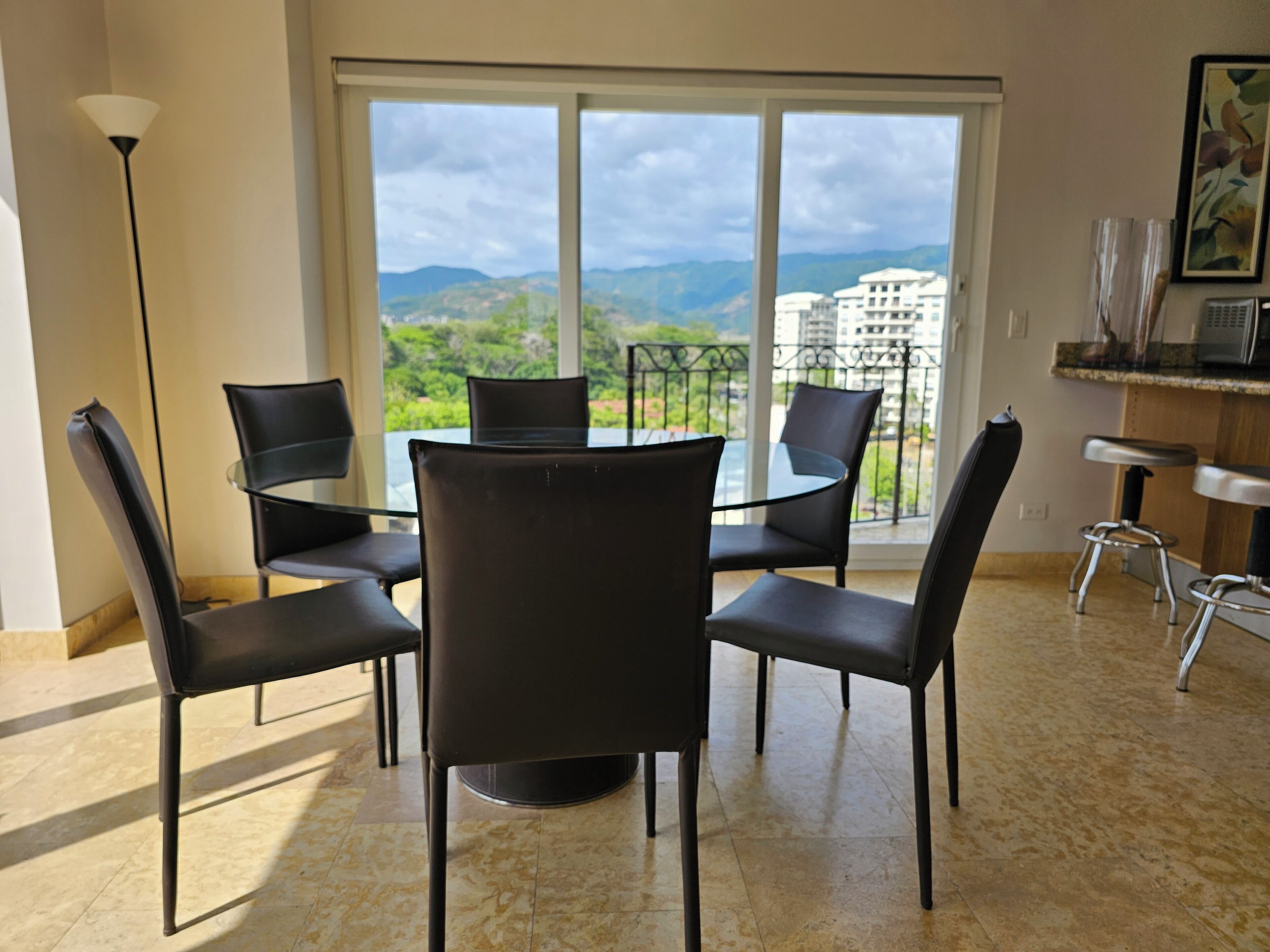 A dining area with a table and chairs, a view of greenery and buildings through large windows, and a kitchen area with a counter and stools.