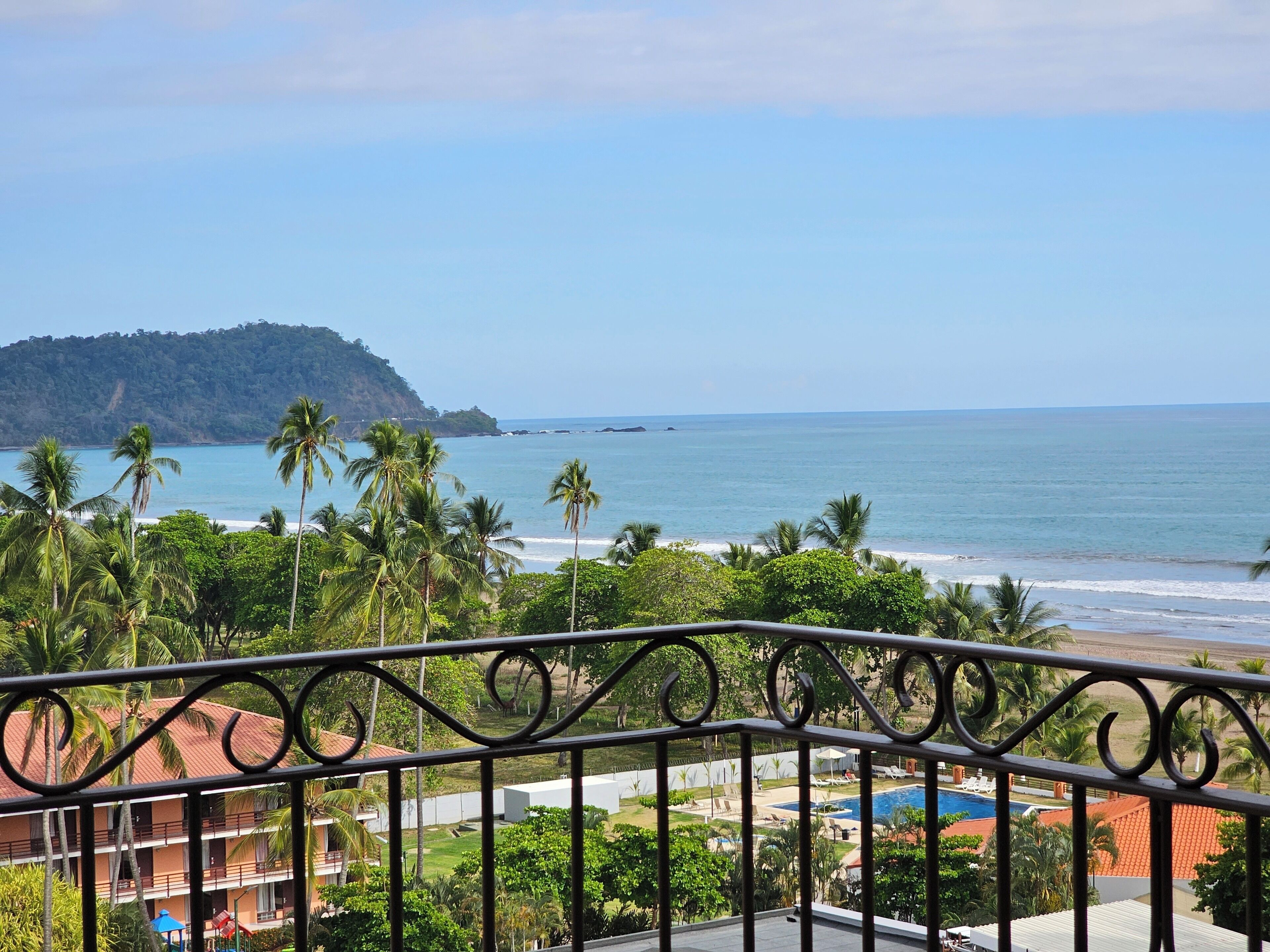 Un balcón con vista a una playa, palmeras y montañas al fondo.