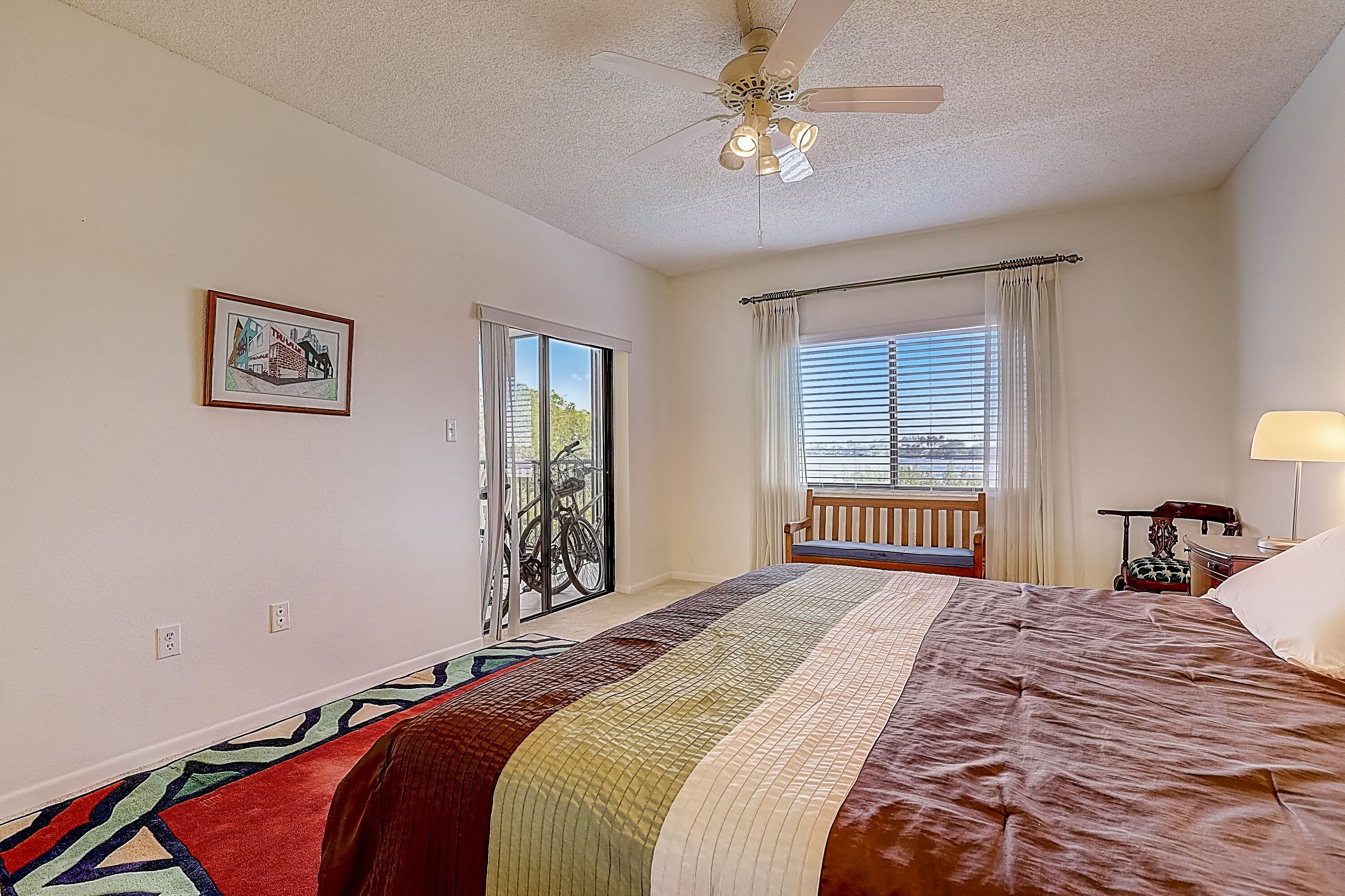 Ein Schlafzimmer mit einem großen Bett, einem Deckenventilator, einem Fenster mit Vorhängen, einer Tür, die auf einen Balkon führt, und einem gerahmten Bild an der Wand.