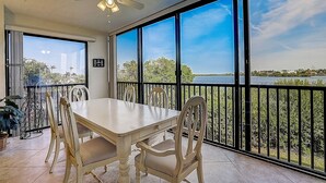 A dining area with a table and chairs, large windows with a view of a body of water and greenery, and a ceiling fan.