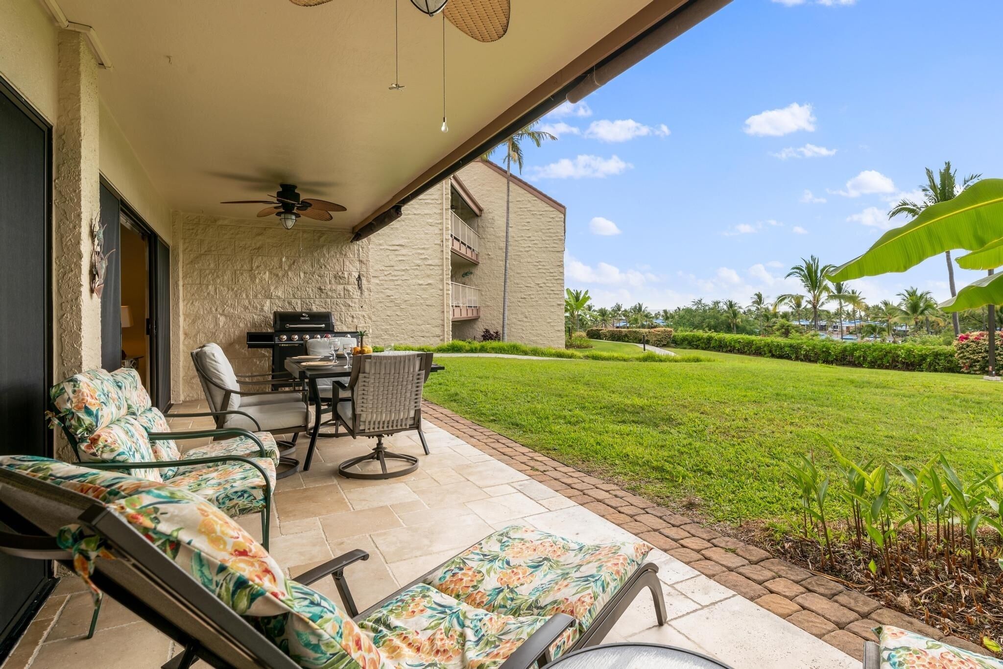 An outdoor patio with a dining table and chairs, a ceiling fan, and a view of a well-maintained lawn and palm trees.