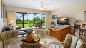 A spacious living area with a glass dining table, wicker chairs, a TV, and a large window overlooking a lush garden.