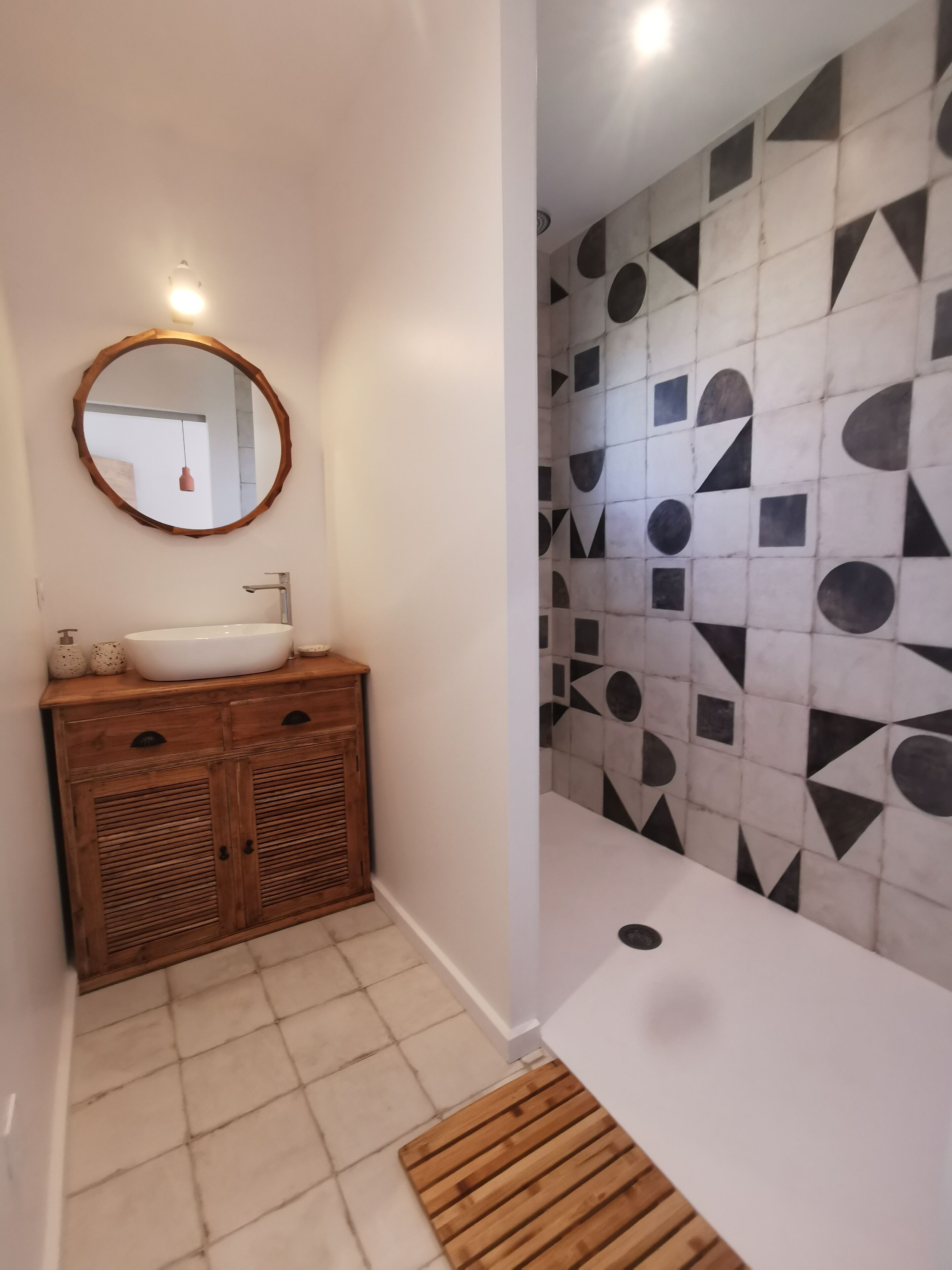 A bathroom with a wooden vanity, a round mirror, and a tiled wall with geometric patterns.