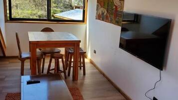 Una habitación con una mesa de comedor, sillas, un televisor montado en la pared y un ventanal con vistas a árboles.