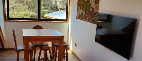 A room with a dining table, chairs, a TV mounted on the wall, and a large window with a view of trees.