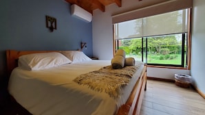 A bedroom with a wooden ceiling, a bed with white linens, a window with a view of greenery, and a small nightstand with a lamp.