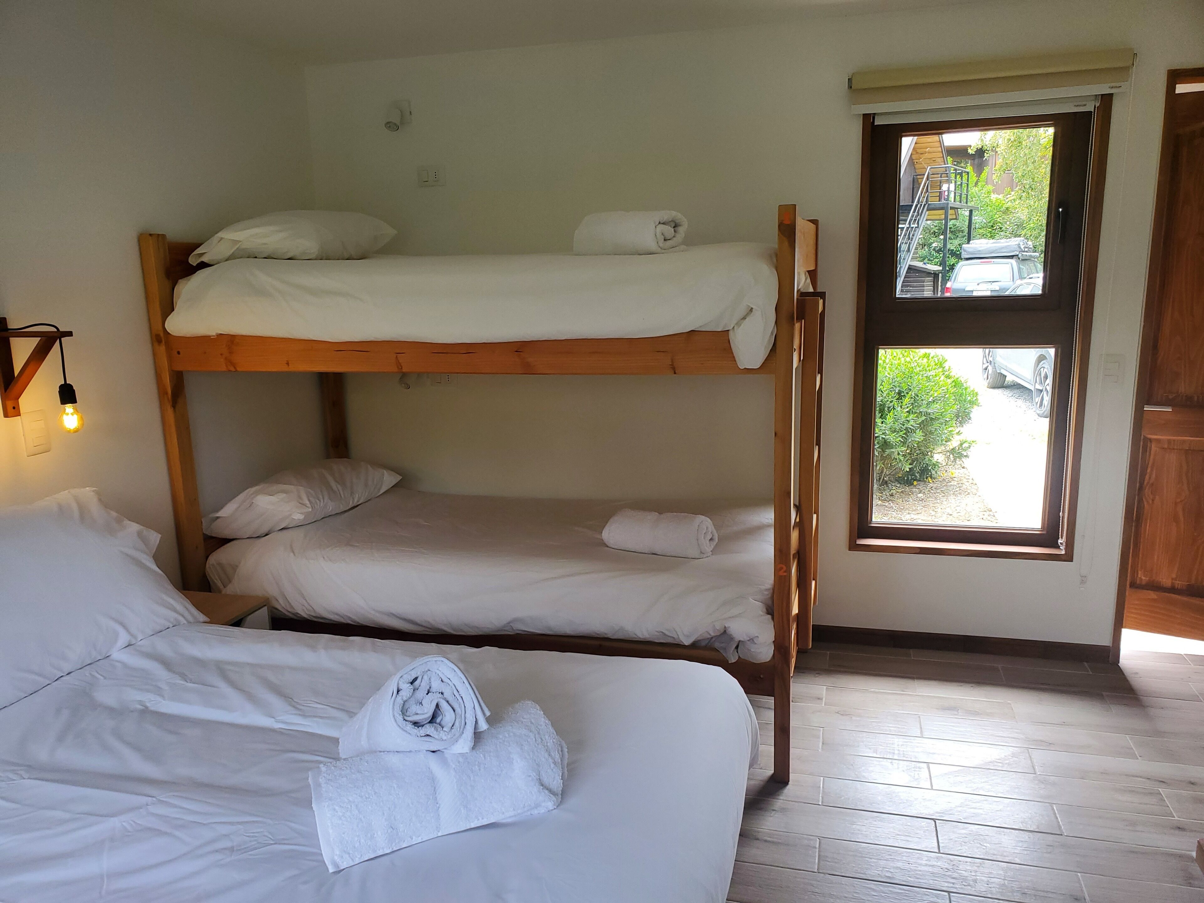 A bunk bed room with two beds, a window with a view of greenery and a parked car, and a wall-mounted light fixture.