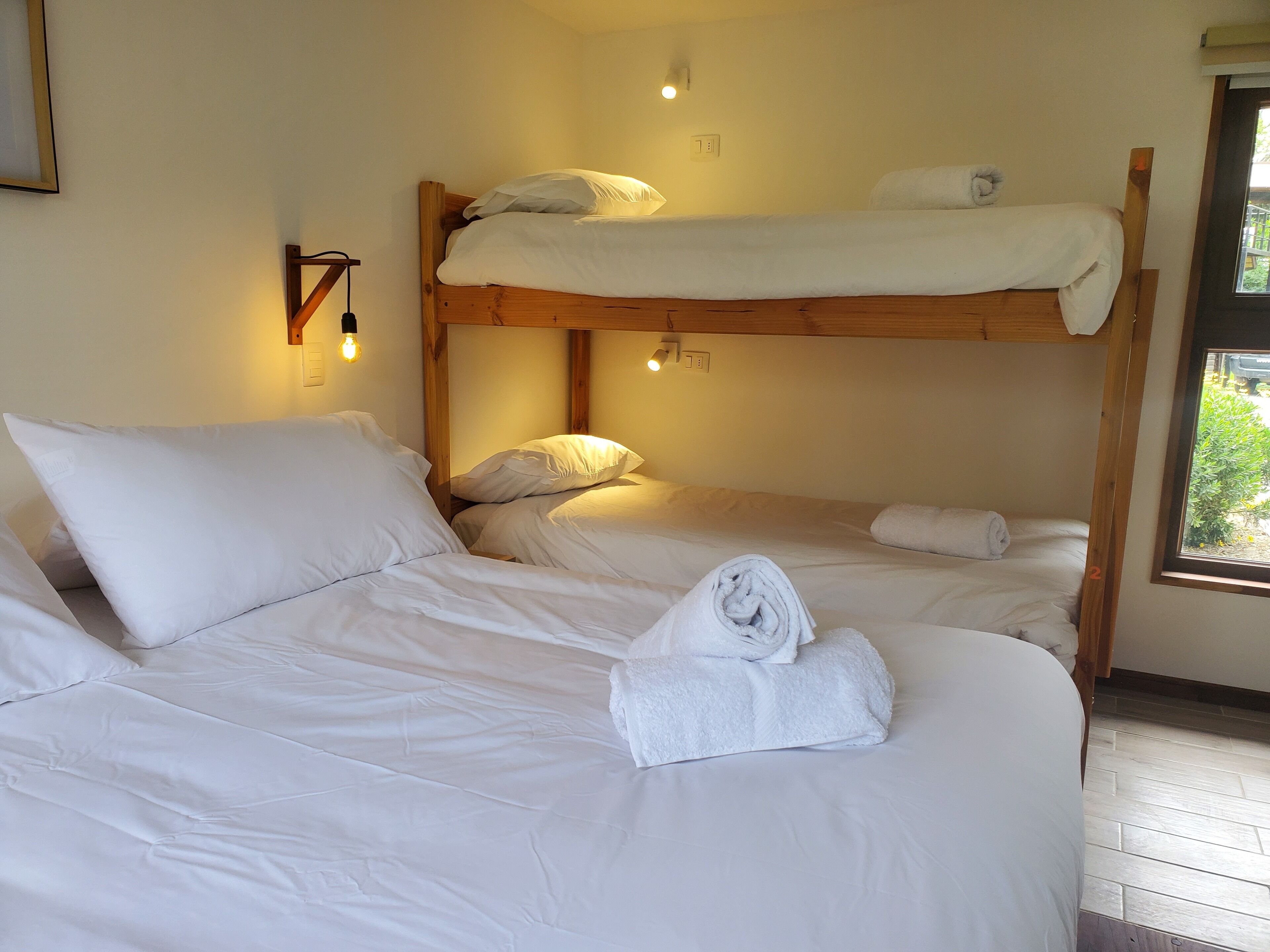 Een slaapkamer met stapelbed, witte beddengoed, een houten hoofdbord en een raam met uitzicht op groen.